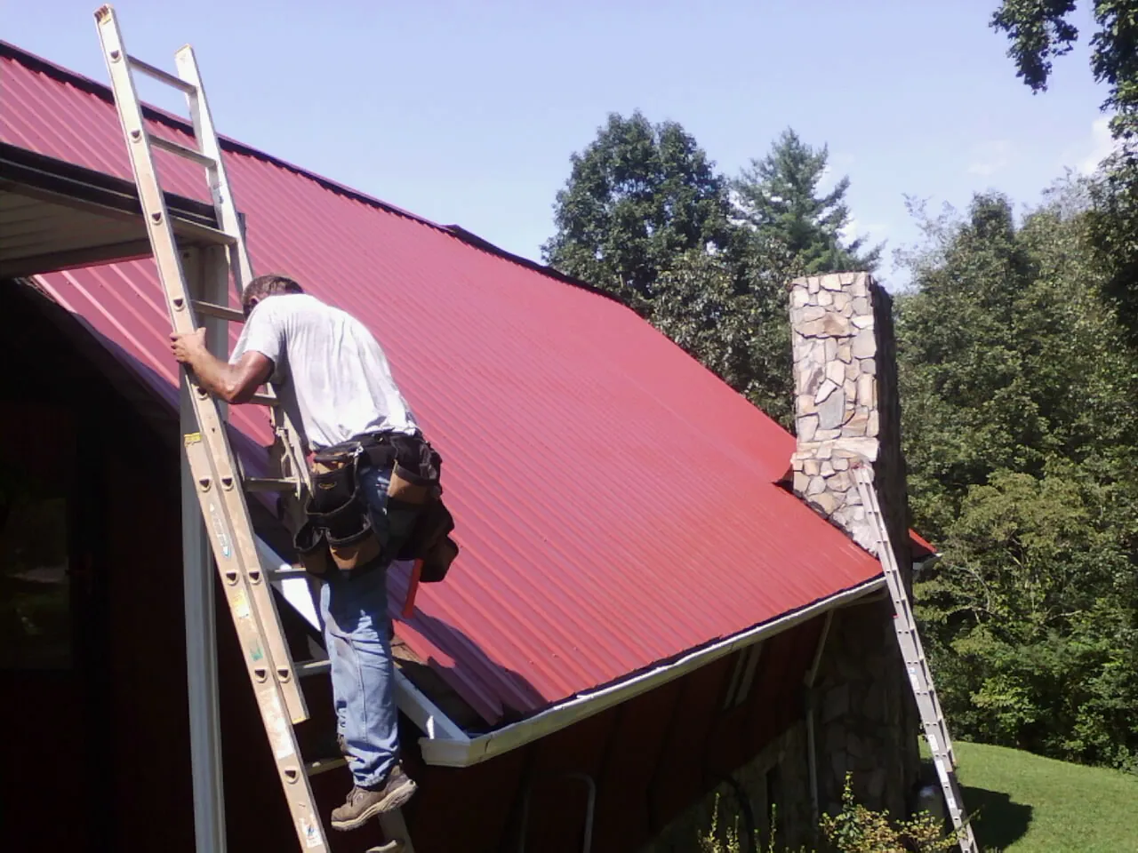 Roofer on a ladder beside a red metal roof and stone chimney — Soffit & Fascia Repair by Roofing Brothers in Jellico